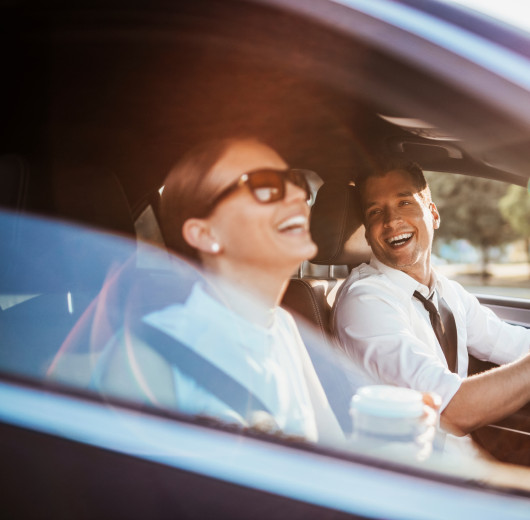 Mann und Frau mit weißem Hemd sitzen lachend in einem schwarzen Auto während die Sonne hinein scheint.