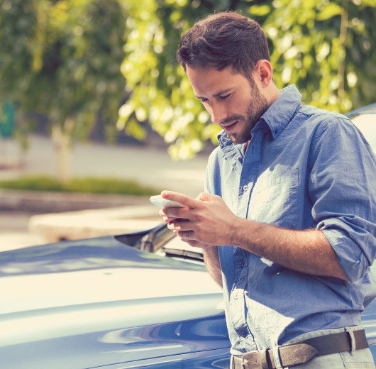 Mann im blauen Hemd steht vor einem blauen Auto und schaut auf sein Handy.