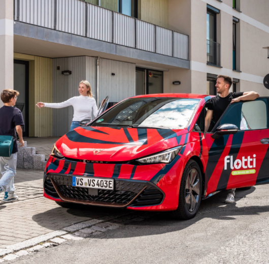 Rotes Elektroauto mit schwarzem Muster und flotti-Logo, geparkt vor einem Wohngebäude, Mann, Frau und Kind drumherum.