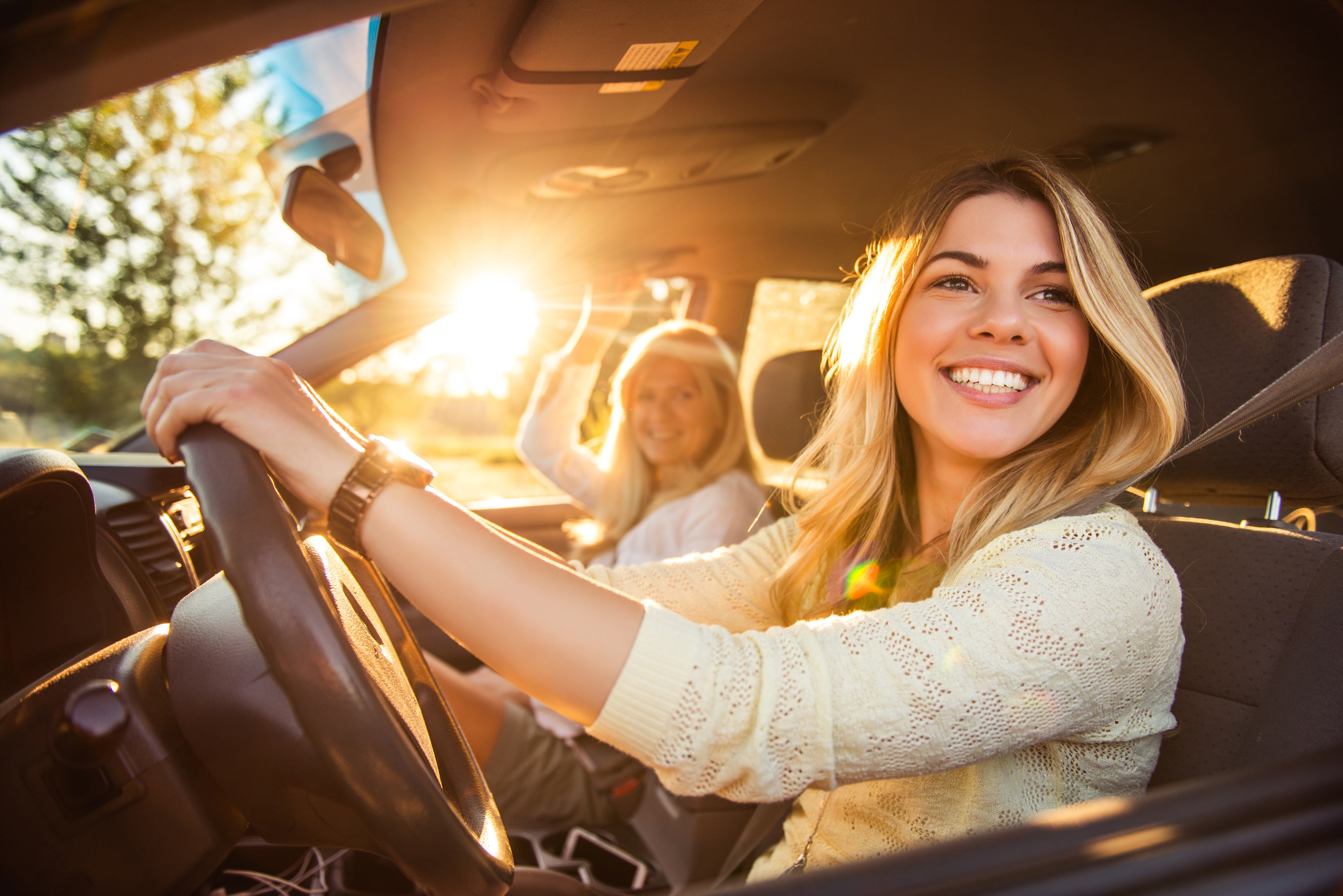 Zwei Frauen sitzen lächelnd in einem Auto, eine hält das Lenkrad, Sonnenlicht fällt durch die Seitenscheibe. ?>