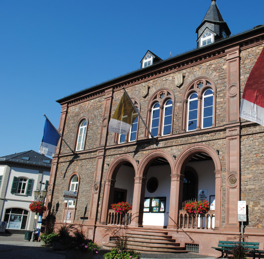 Historisches Gebäude aus rotem Sandstein mit drei Rundbögen, Fahnen und Blumenschmuck vor blauem Himmel.
