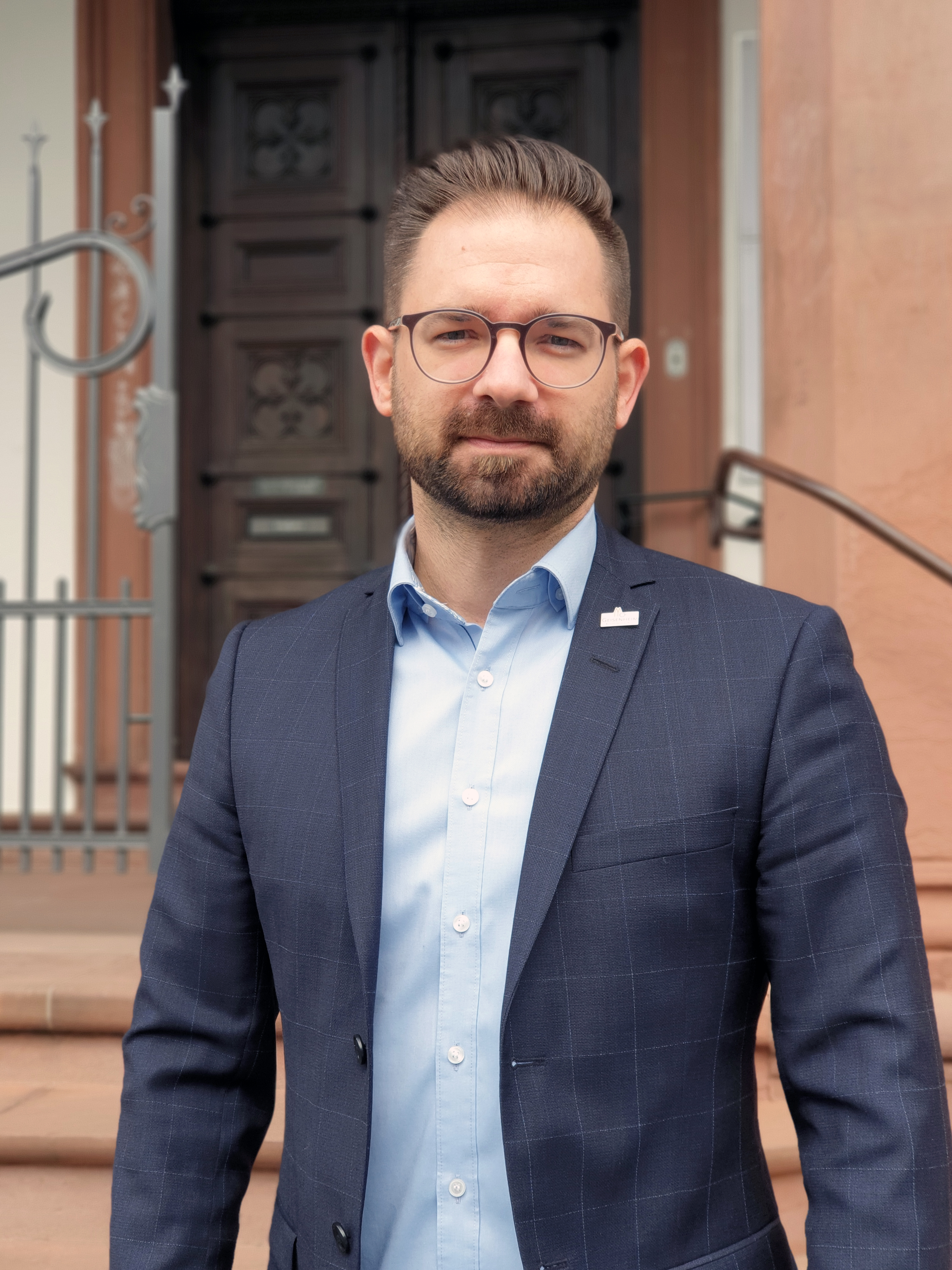 Person in dunkelblauem Anzug, hellblauem Hemd und Brille steht vor historischem Gebäude mit Holztür. ?>