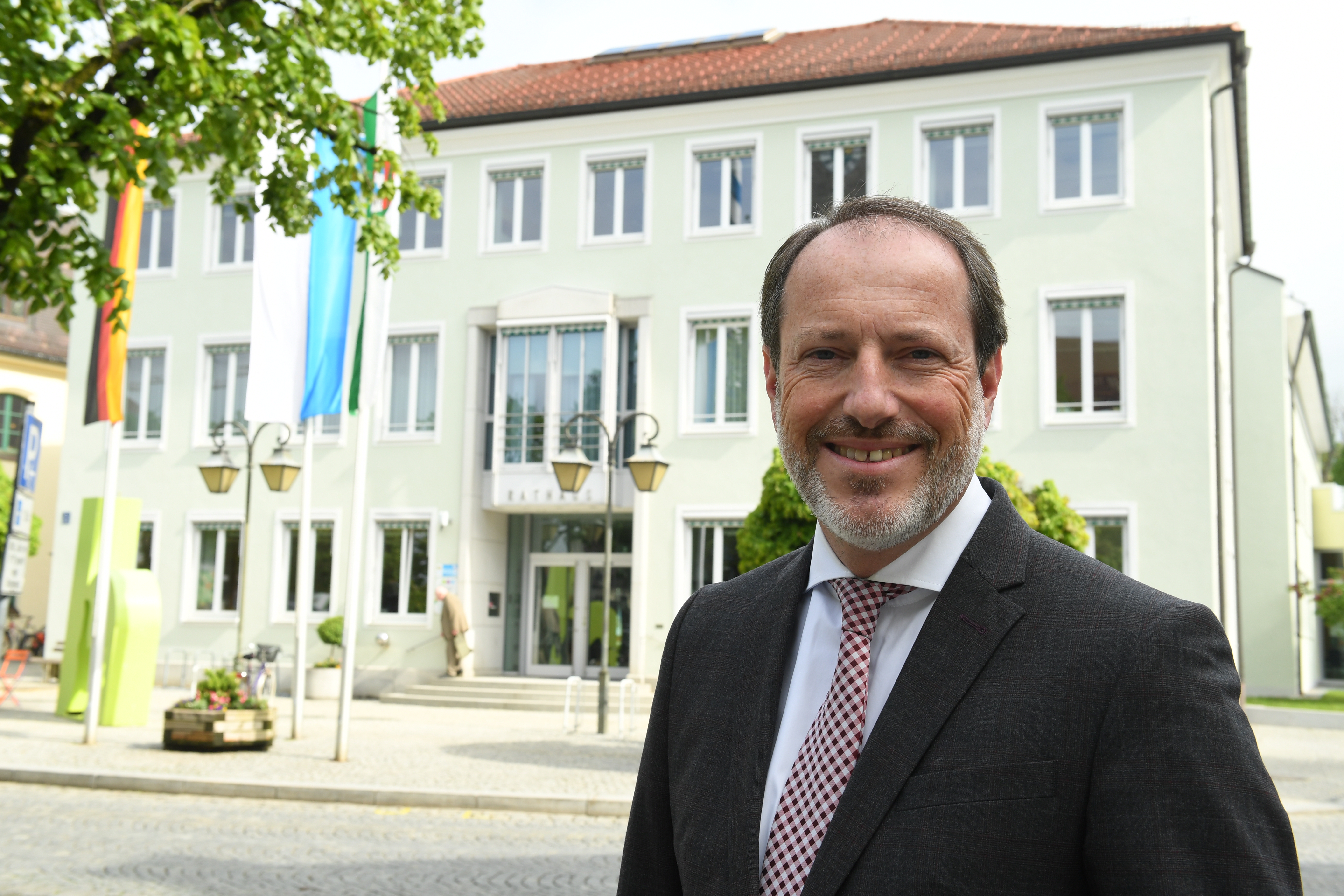 Person im Anzug steht vor einem Rathaus mit Fahnen und hellem Gebäude im Hintergrund ?>