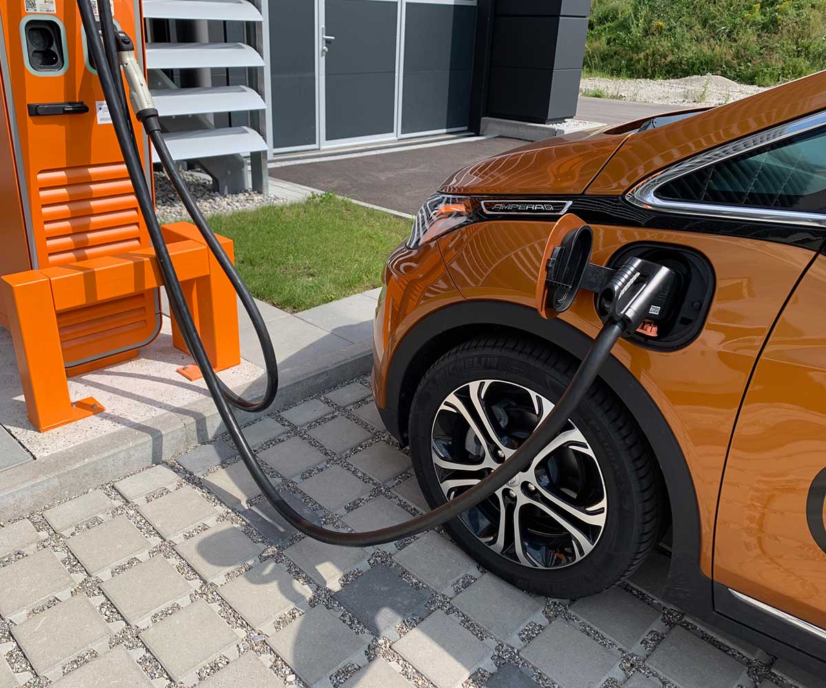 Oranges Elektroauto wird an einer Ladestation mit schwarzem Kabel geladen, im Hintergrund modernes Gebäude. ?>