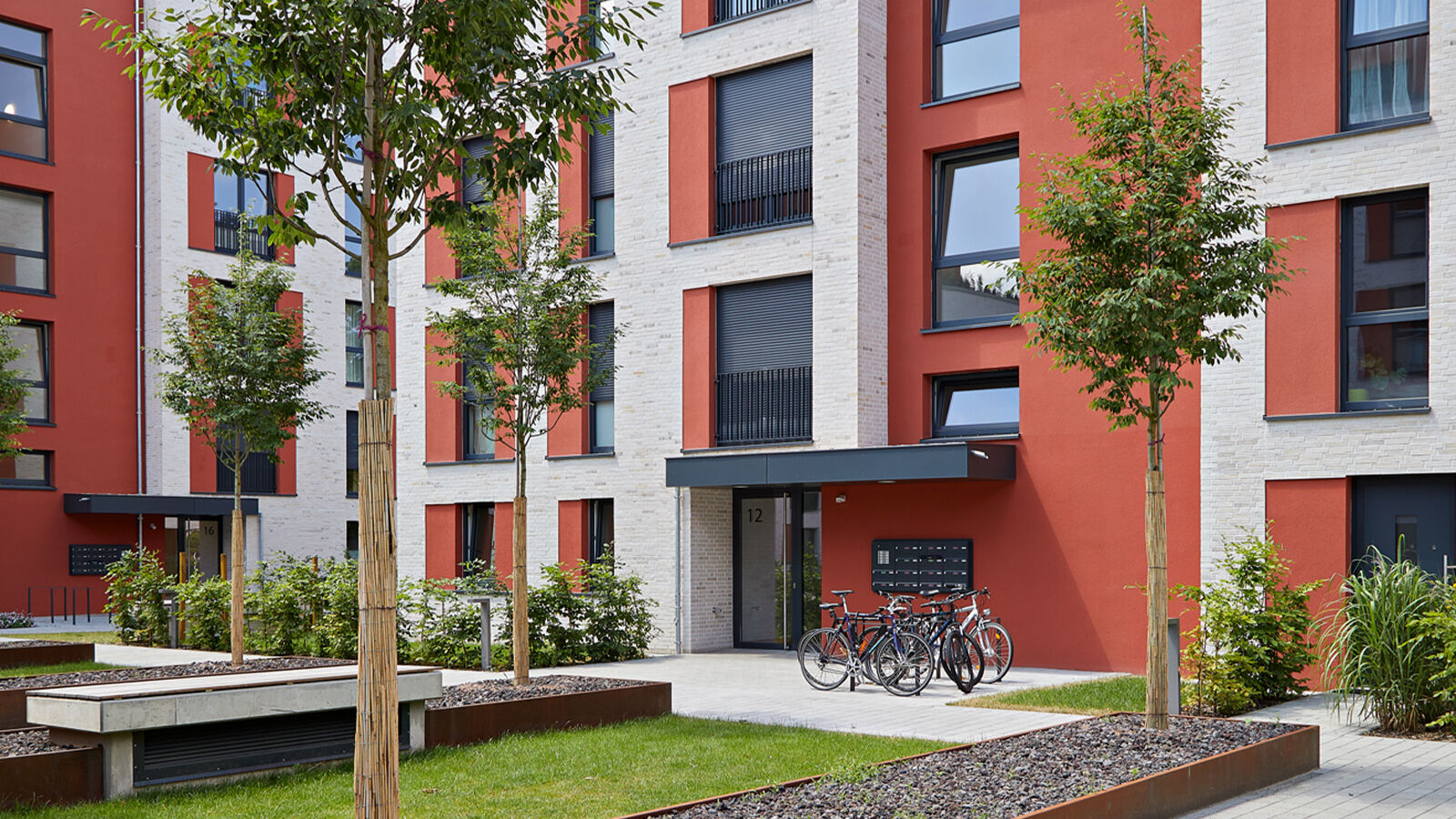 Moderner Eingangsbereich eines Wohngebäudes mit roten und weißen Fassaden, Grünflächen, Bäumen und abgestellten Fahrrädern. ?>