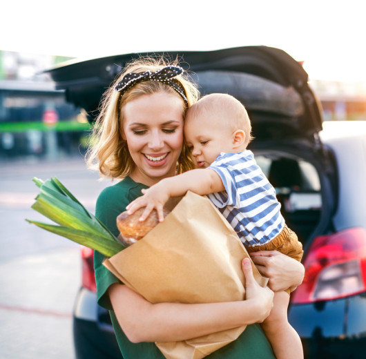 Frau mit Kind und Einkaufstüte im Arm steht vor schwarzem Auto.