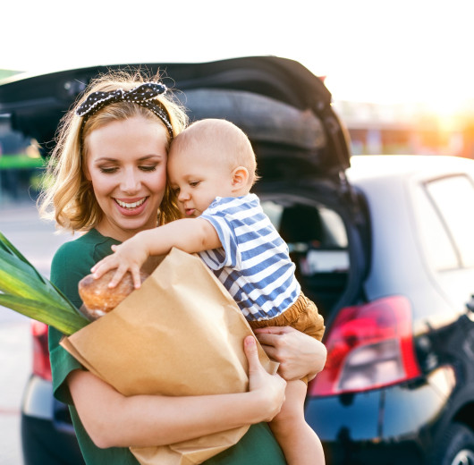 Frau mit Kind und Einkaufstüte im Arm steht vor schwarzem Auto.