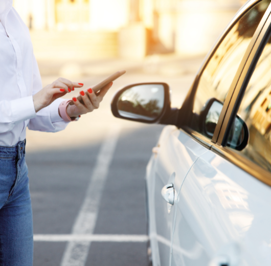 Frau mit weißem Hemd und Jeans bedient Smartphone neben weißem Auto auf Parkplatz.