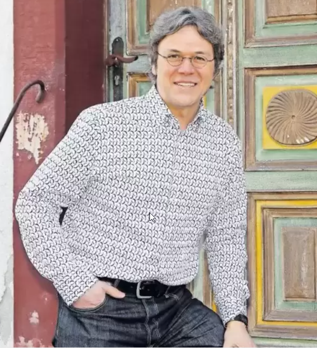 Mann in gemustertem Hemd und Jeans steht vor einer dekorativen, bunt gestrichenen Holztür. ?>