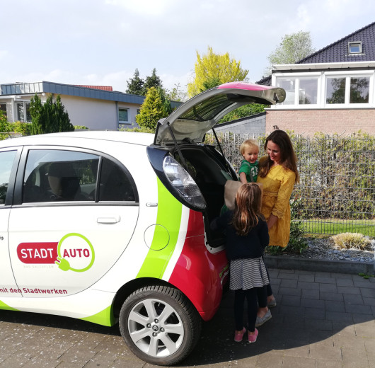Person lädt Gepäck in den Kofferraum eines StadtAuto-Fahrzeugs mit grün-rotem Design, zwei Kinder stehen daneben.