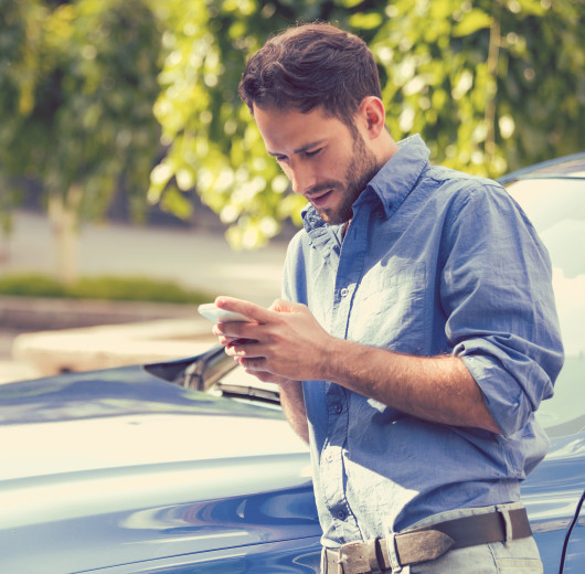 Mann im blauen Hemd steht vor einem blauen Auto und schaut auf sein Handy.