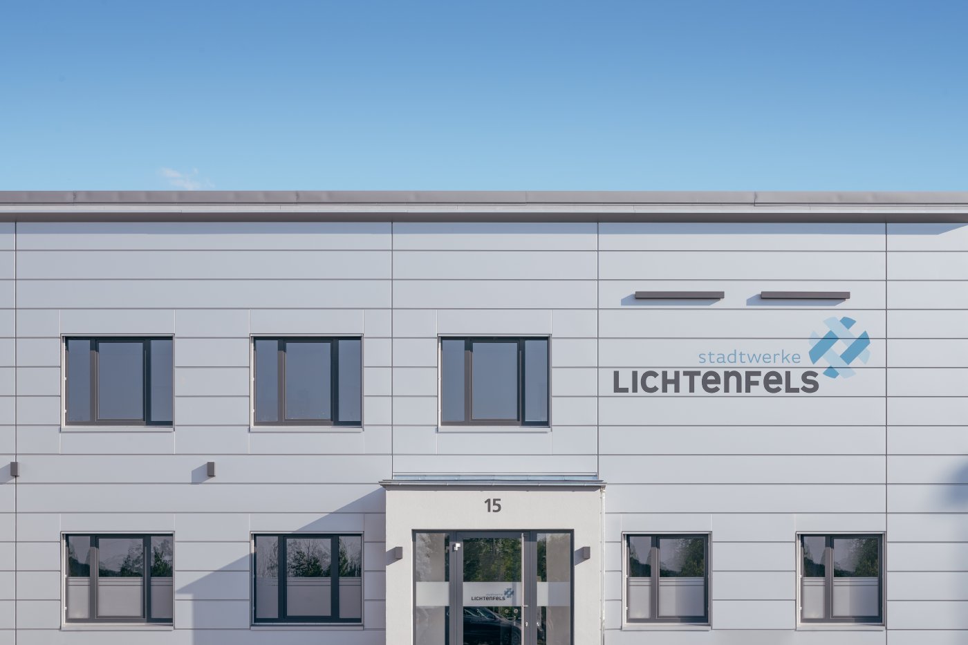 Bürogebäude mit weißer Fassade, einigen Fenstern und Logo ‚Stadtwerke Lichtenfels‘ auf der Vorderseite unter blauem Himmel. ?>