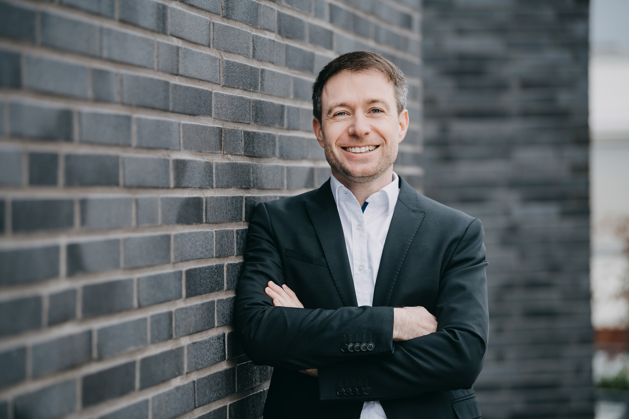 Person in schwarzem Anzug und weißem Hemd steht mit verschränkten Armen vor einer modernen Backsteinwand. ?>