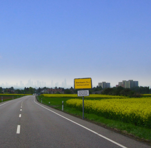 Straße mit Ortsschild Steinbach (Taunus) vor gelbem Rapsfeld, im Hintergrund Skyline von Frankfurt am Main.