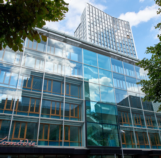 Verglastes Bürogebäude, mit Hochhaus dahinter, links und recht grüne Baumkronen.