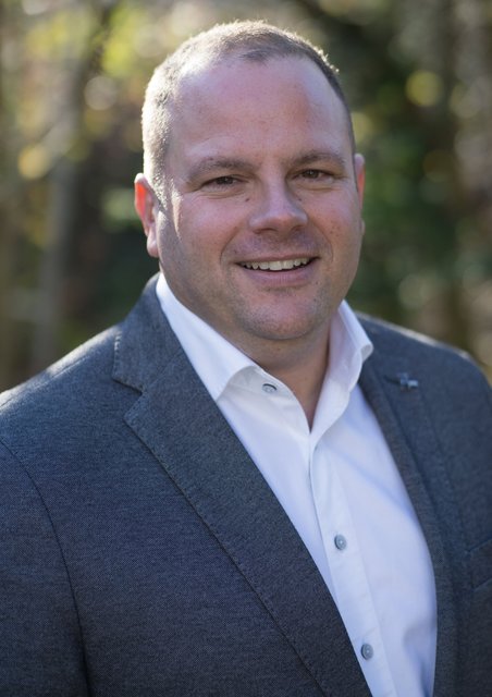 Person in grauem Sakko und weißem Hemd steht im Freien vor unscharfem Hintergrund mit Bäumen. ?>