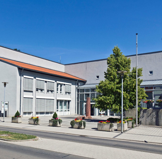 Verwaltungsgebäude mit grauer Fassade, großen Fenstern, Pflanzkübeln und Fahnenmasten an einem gepflasterten Platz.