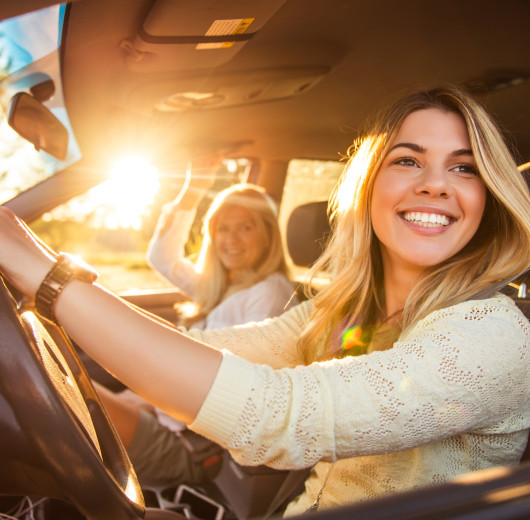 Zwei Frauen sitzen lächelnd in einem Auto, eine hält das Lenkrad, Sonnenlicht fällt durch die Seitenscheibe.