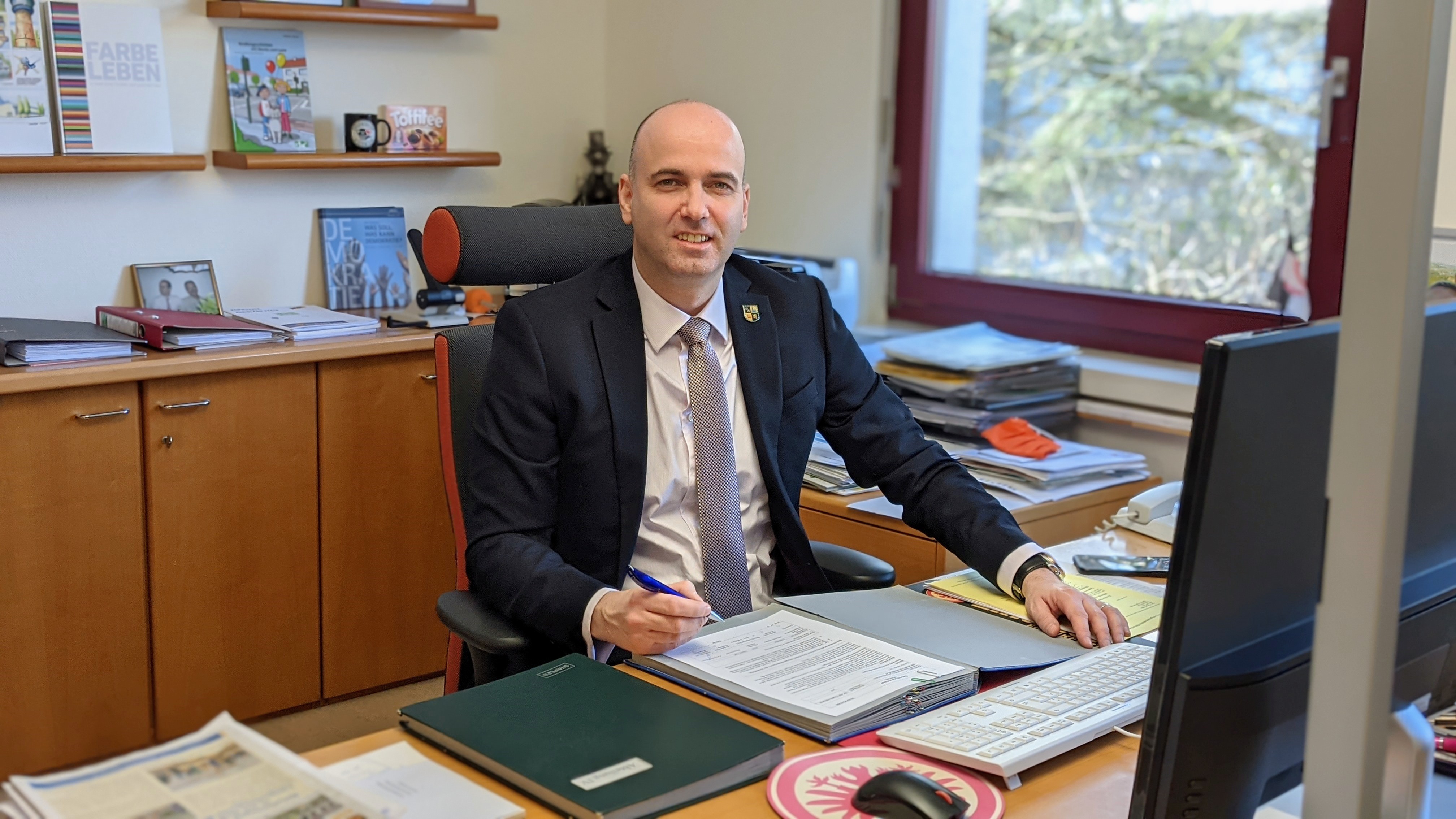 Michael Cyfka sitzt an einem Schreibtisch im Büro, vor sich ein Computerbildschirm und Aktenordner. ?>